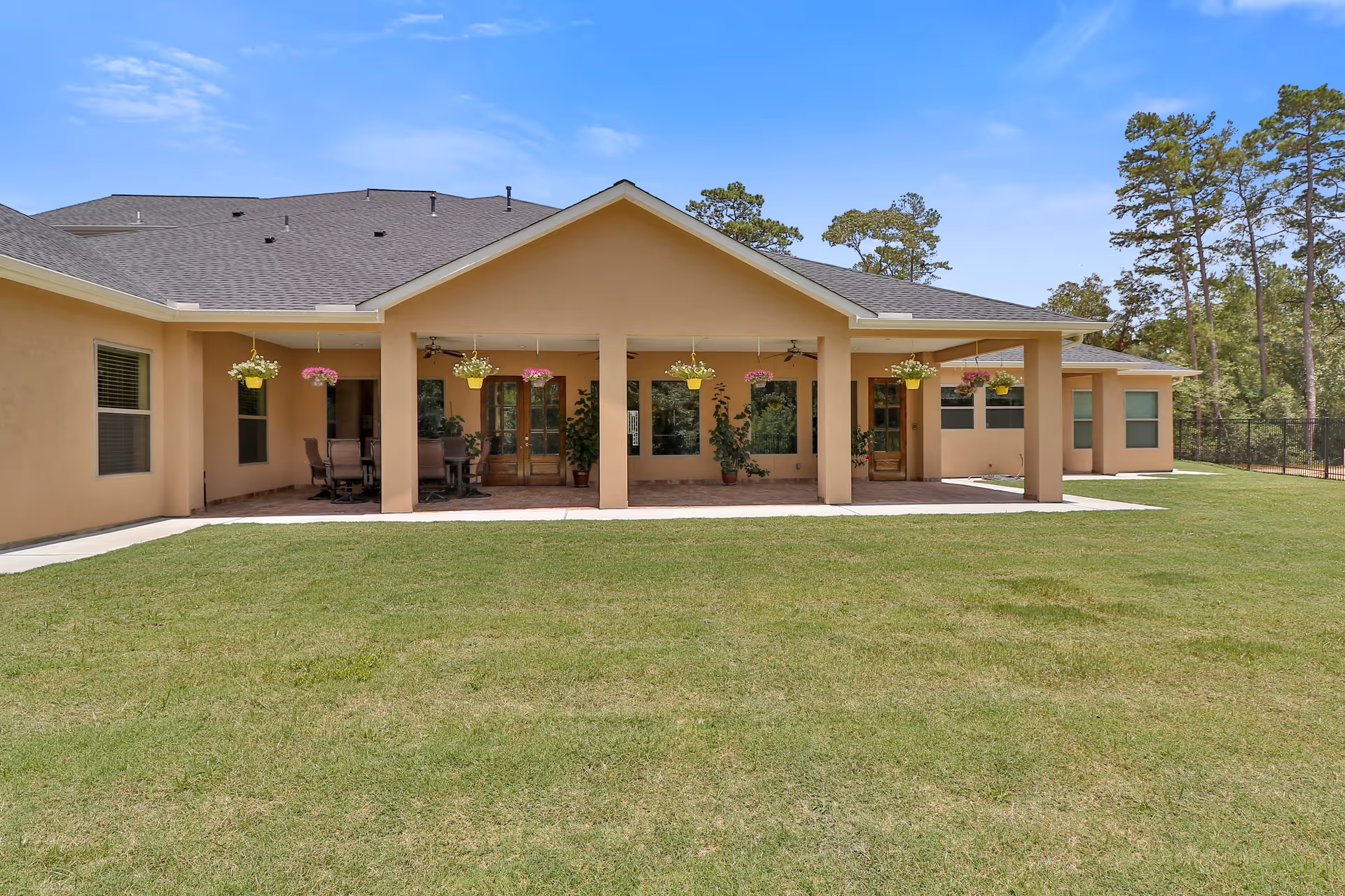 Exterior view of a single-story building with a covered patio area featuring hanging flower pots and outdoor seating. The building is surrounded by a well-maintained grassy lawn and trees in the background under a clear blue sky.