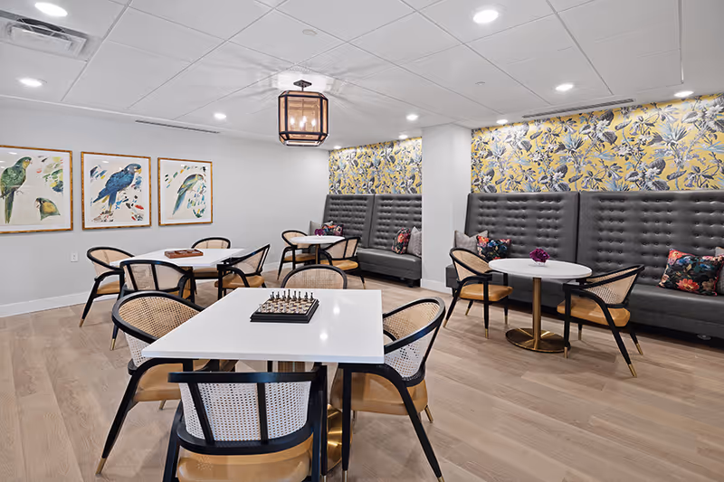 A cozy common area with multiple white tables and black chairs with cane backs. Some tables have chessboards and small flower arrangements. The walls feature colorful bird artwork and a floral yellow wallpaper behind gray tufted bench seating with decorative pillows. The room has light wood flooring and recessed ceiling lights.