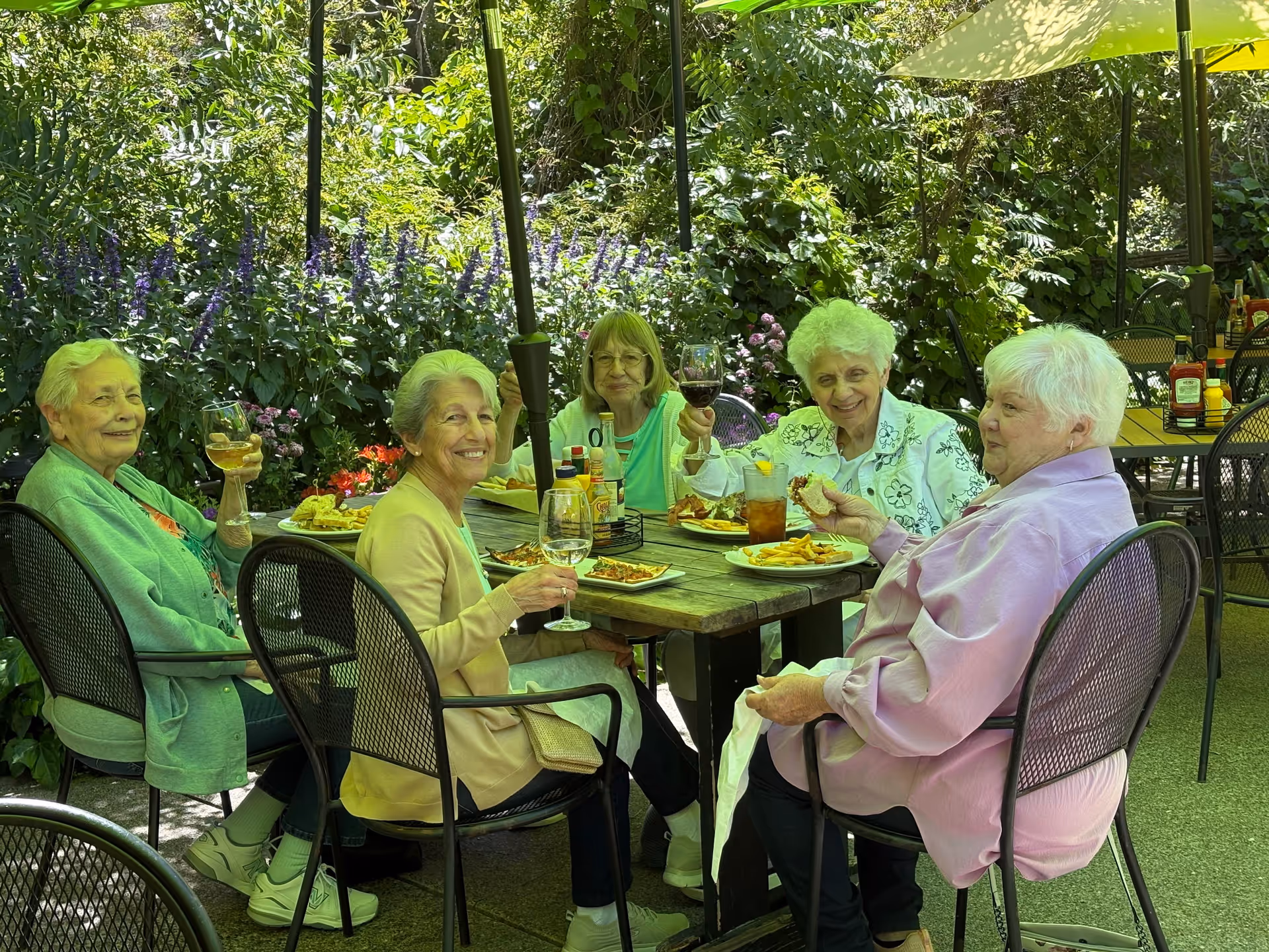 Five elderly women sitting around a wooden outdoor table enjoying a meal together. They are surrounded by lush greenery and flowers, with drinks and plates of food on the table. The women are smiling and appear to be having a pleasant time in a garden-like setting.