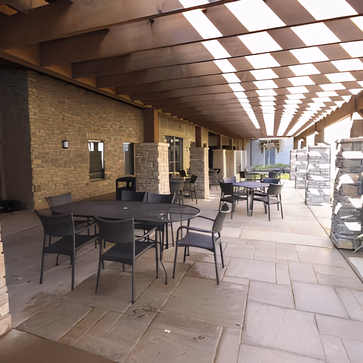 Outdoor covered patio area with multiple black metal tables and chairs arranged on a stone tile floor. The ceiling has a patterned design allowing sunlight to filter through, casting shadows on the floor. Stone pillars and walls line the left side, with large windows and doors visible.
