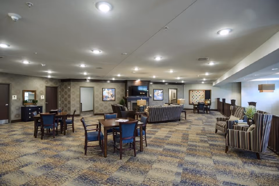 Spacious furnished common room with several dining tables, upholstered chairs and sofas arranged around a fireplace and TV.