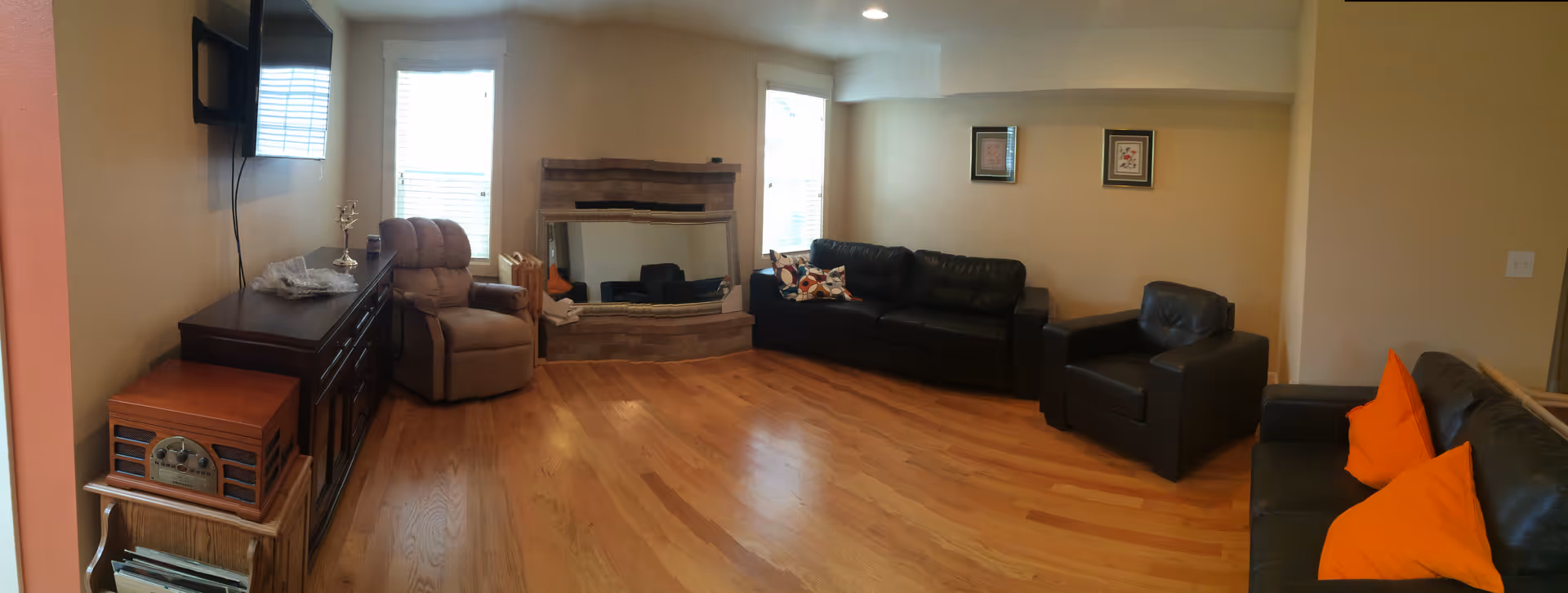 A living room with wooden flooring, featuring a brown recliner chair, a dark wooden cabinet with a vintage radio on a small side table, a wall-mounted TV, a stone fireplace between two windows, and three black leather sofas with orange and patterned cushions.