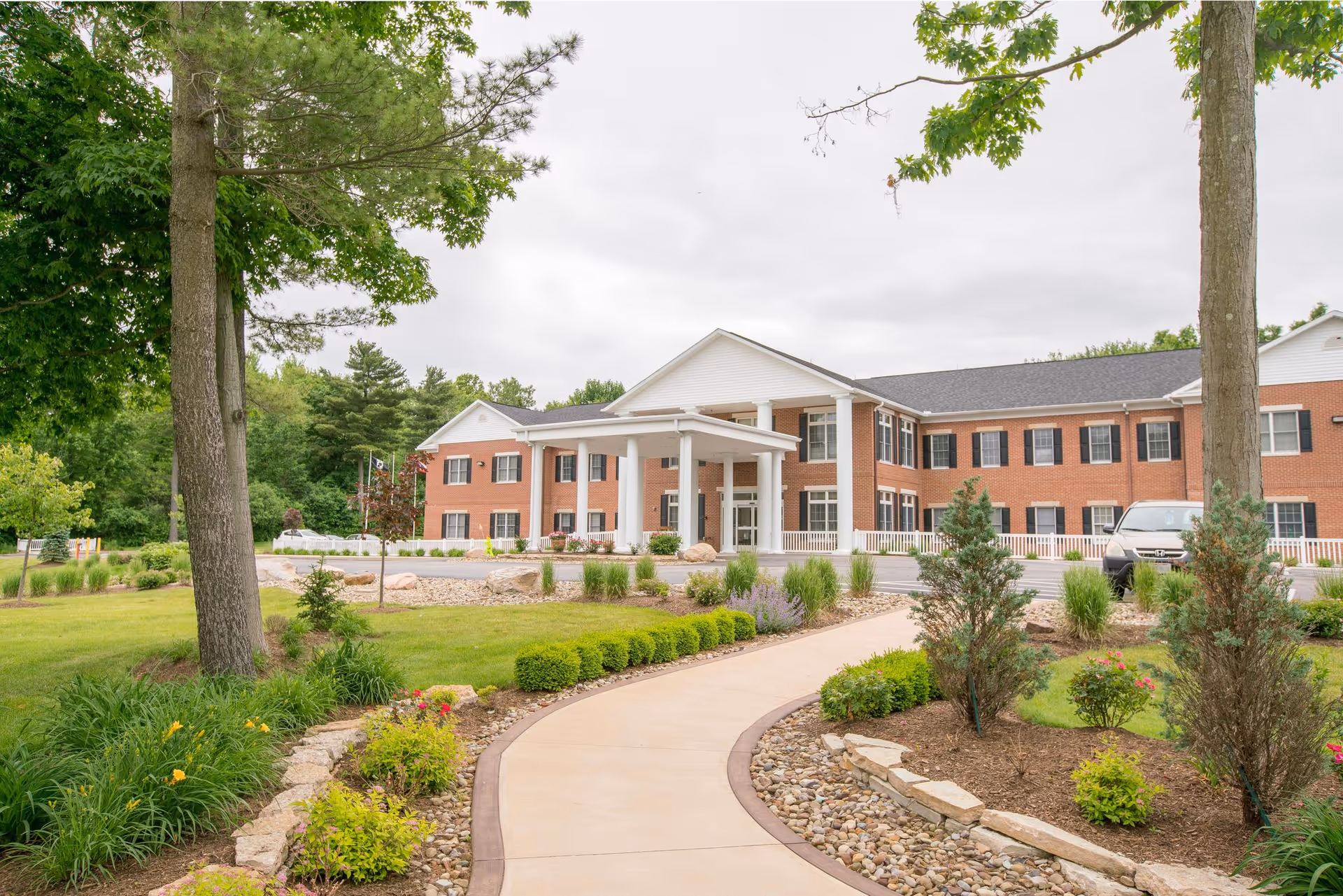 Exterior view of a two-story brick building with white columns at the entrance, surrounded by landscaped gardens with trees, shrubs, and a curved walkway leading to the entrance under a cloudy sky.