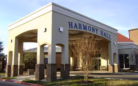 Exterior view of the entrance to Harmony Hall, a building with a covered drop-off area supported by columns, a tree in front, and a clear blue sky.