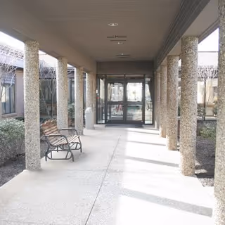 Covered walkway with textured stone columns and a bench on the left side leading to glass double doors at the entrance of a building, with some landscaping visible on the sides.