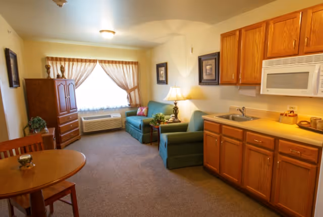 Small senior living studio with a kitchenette and wooden cabinets, a round dining table, green sofa and armchair, and a curtained window.