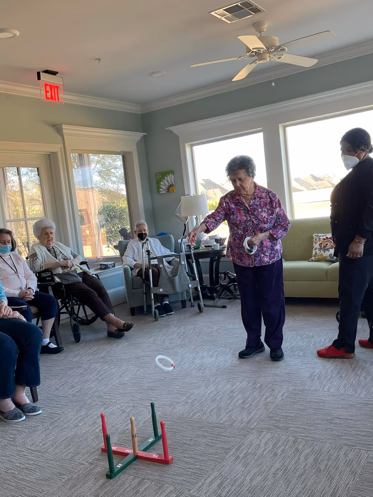 A group of elderly women, some seated and some standing, are playing a ring toss game in a well-lit room with large windows. One woman in a floral shirt is actively tossing a ring towards the wooden ring toss stand on the carpeted floor. Another woman stands nearby watching, and several others sit in chairs and wheelchairs along the wall. The room has light-colored walls, a ceiling fan, and an exit sign above a door.