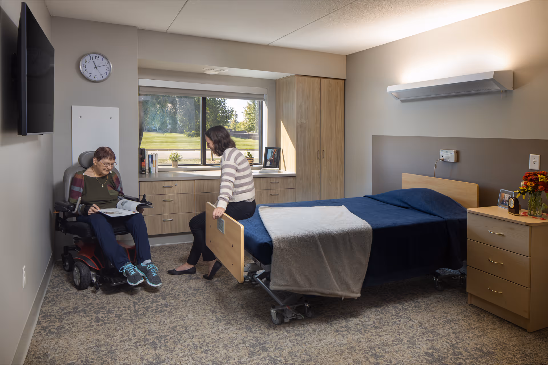 A furnished senior-care private bedroom with a bed, dresser and window, where a woman in a wheelchair and another woman sit and talk.