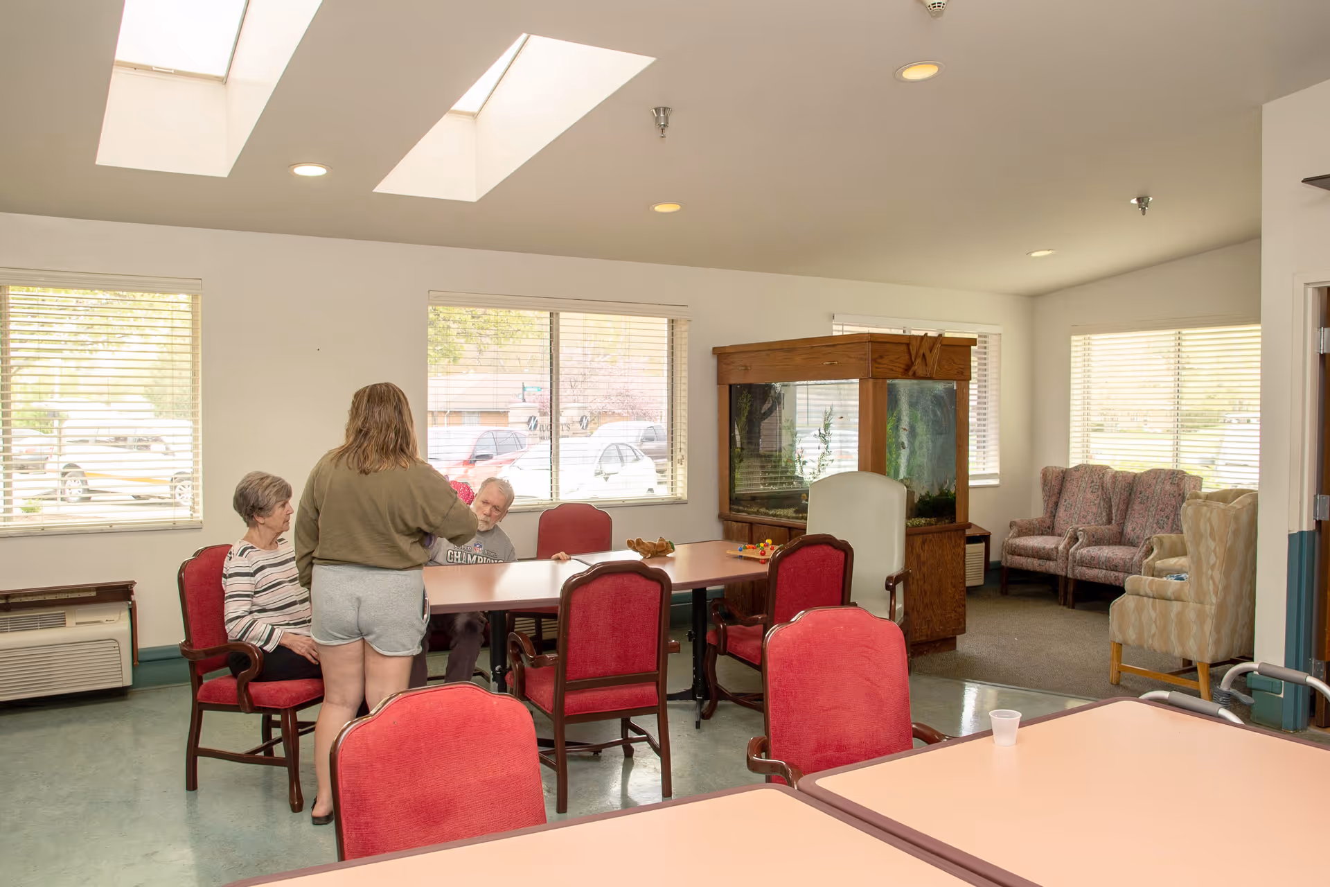 A common area in a senior living facility with several red cushioned chairs around tables. Three people are interacting near one table, with one standing and two seated. The room has large windows with blinds, skylights, and an aquarium in the corner. Additional seating with patterned armchairs is visible in the background.