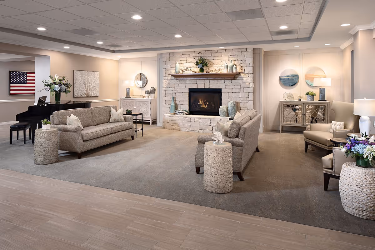 A spacious and elegantly decorated senior living facility common area featuring two beige sofas, a stone fireplace with decorative vases and plants on the mantel, a grand piano with a stool, and various side tables with floral arrangements. The room has soft lighting, neutral tones, and artwork on the walls including an American flag and abstract paintings.