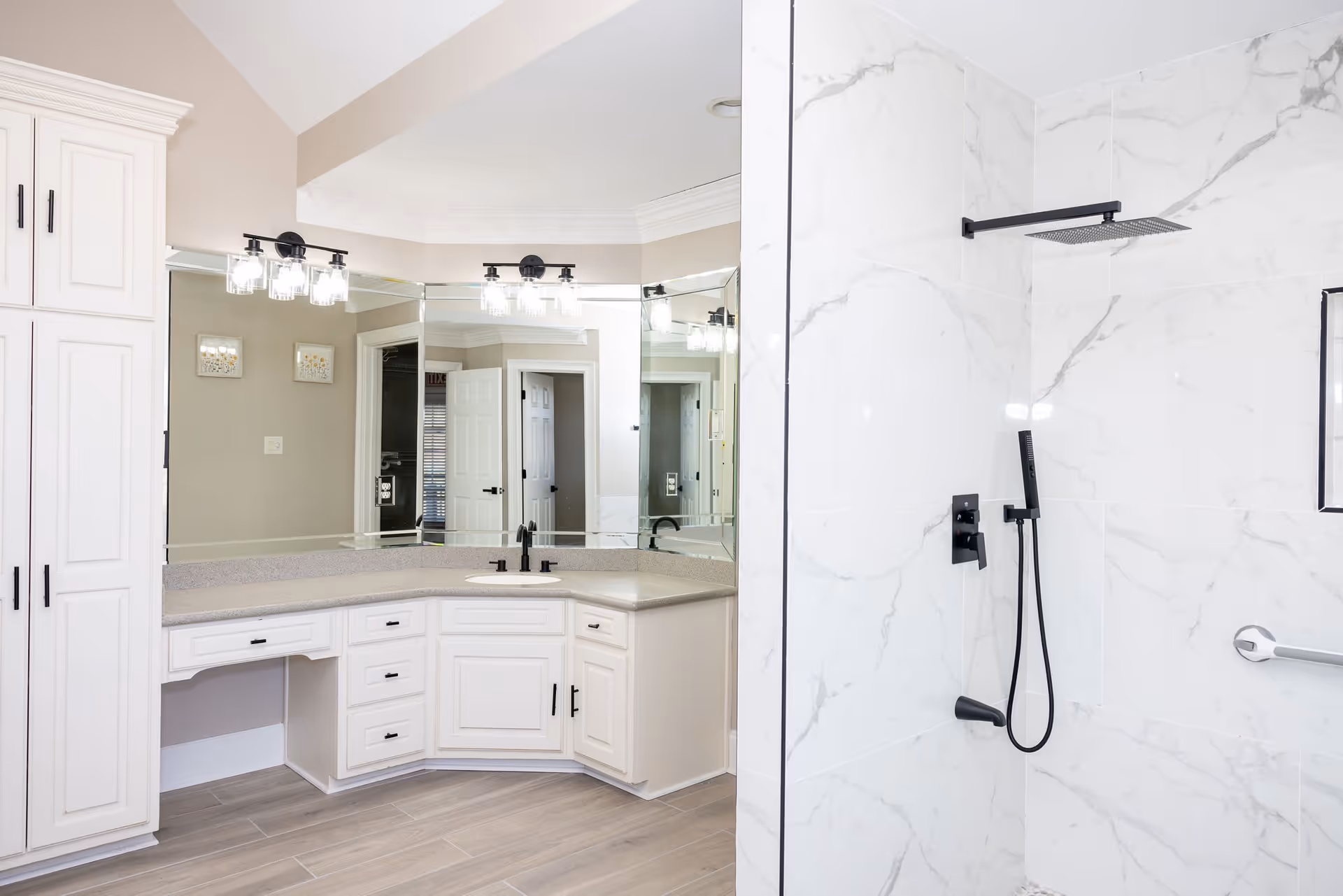 Bright bathroom featuring white cabinetry and a large mirrored vanity next to a marble-tiled walk-in shower with black fixtures.