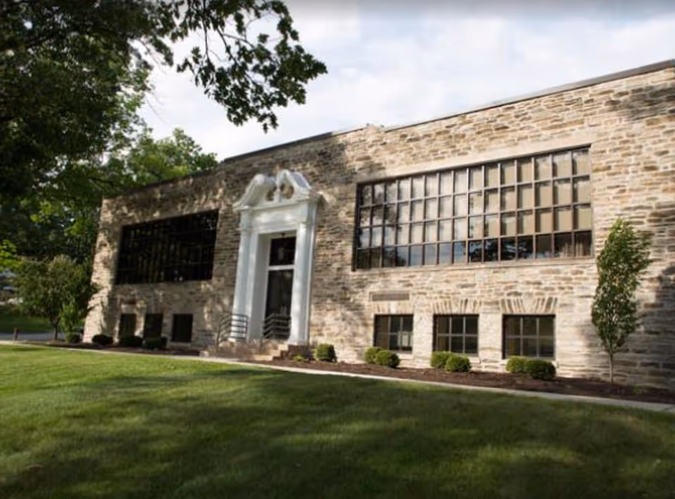 Exterior view of a stone building with large windows and a white decorative entrance surrounded by green grass and trees.
