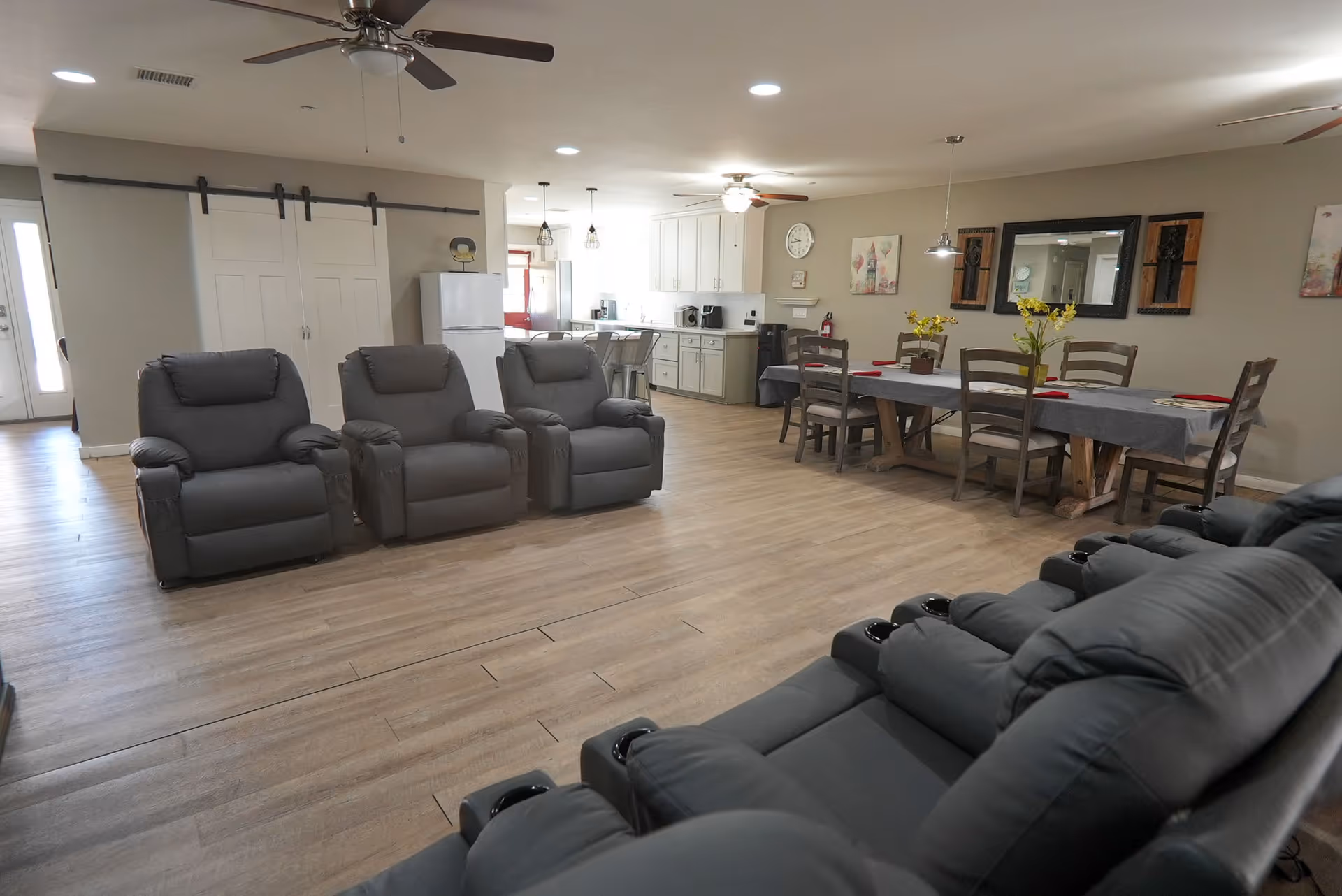 Spacious communal living area with gray recliners, a dining table, and a kitchenette in the background.