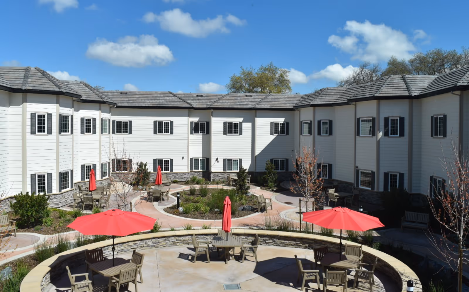 Outdoor courtyard area of a senior living community with circular seating arrangements, tables with red umbrellas, wooden chairs, and landscaped garden beds surrounded by a two-story white building with multiple windows under a blue sky with scattered clouds.