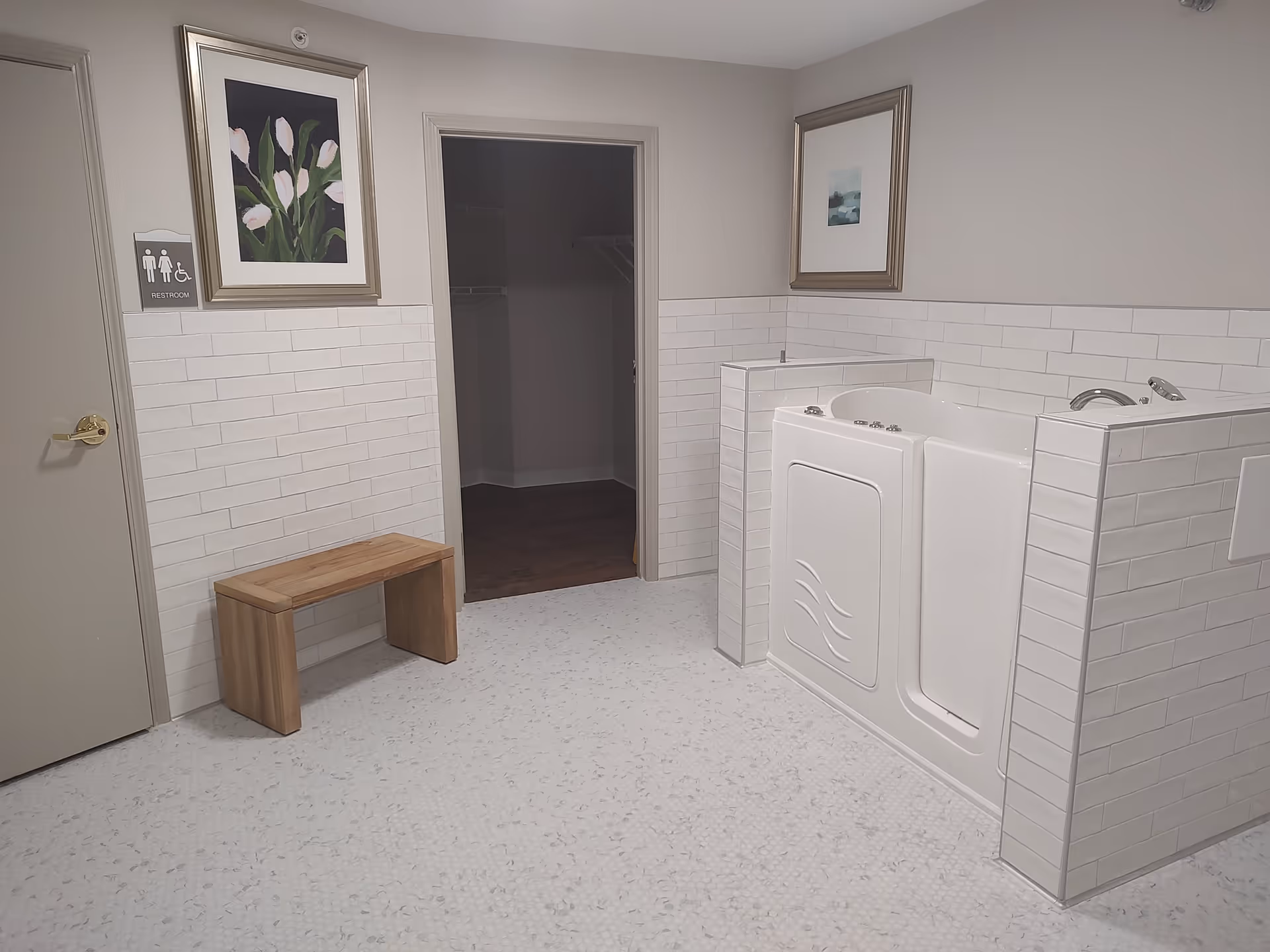 Accessible bathroom area featuring a white walk-in tub, a small wooden bench, framed artwork, and a doorway to a closet.