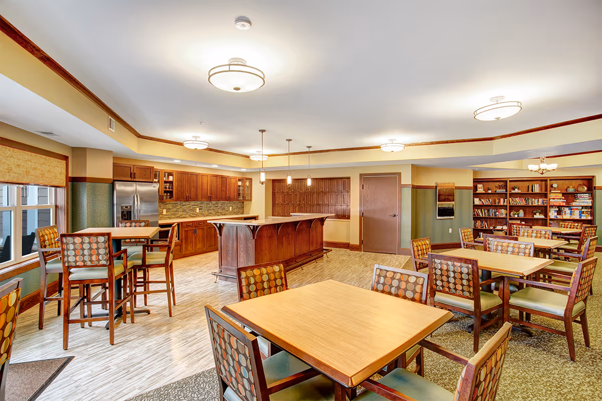 A spacious and well-lit common area in a senior living facility featuring multiple wooden tables and chairs with patterned upholstery. The room includes a kitchen area with wooden cabinets, a stainless steel refrigerator, and a central island with pendant lighting. In the background, there is a bookshelf filled with books and board games, and a door on the far wall.