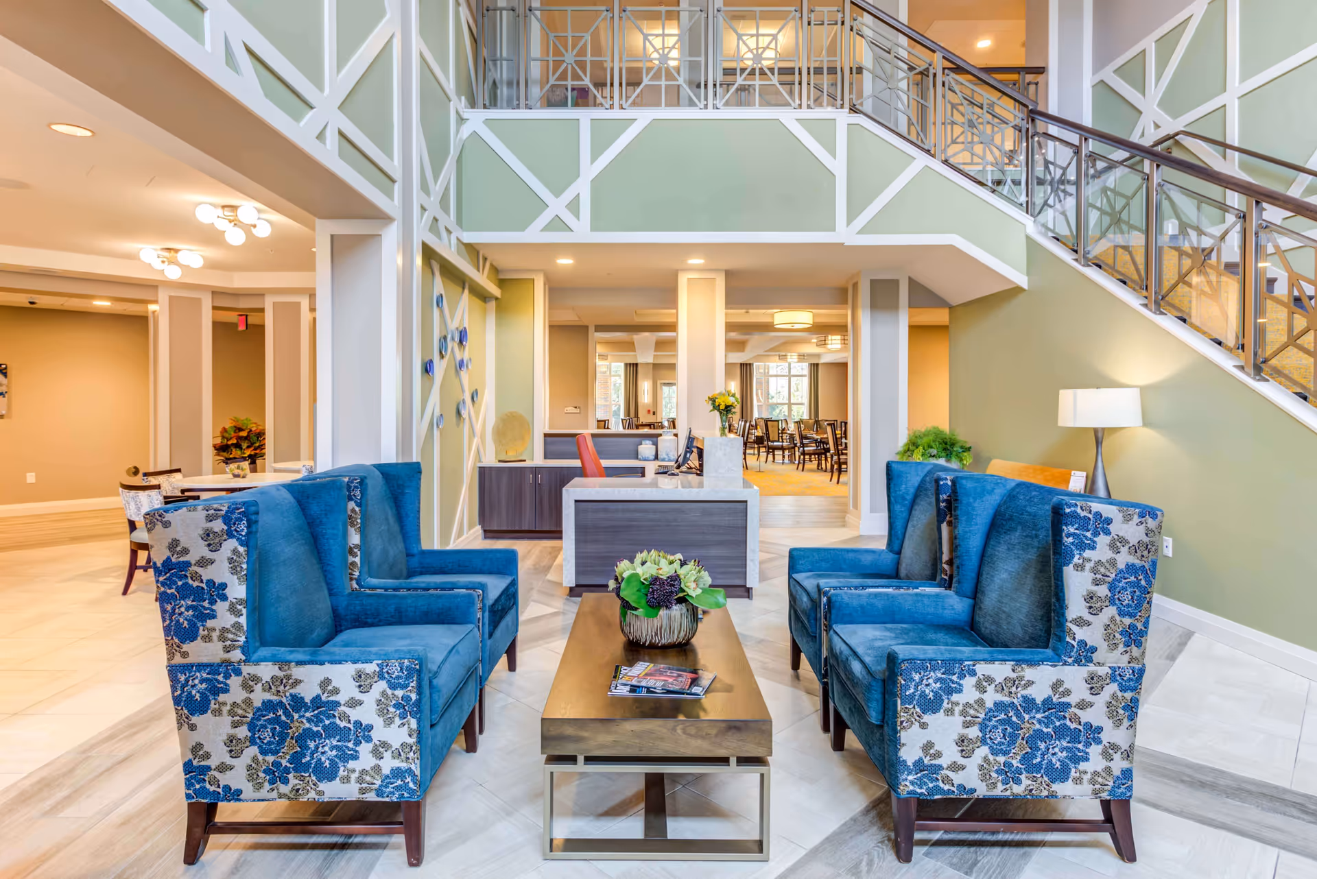 A bright and spacious senior living facility common area with four blue upholstered armchairs featuring floral patterns arranged around a wooden coffee table with a flower arrangement and magazines. In the background, there is a reception desk with a computer and a red chair, and beyond that, a dining area with tables and chairs. The walls are painted green with white trim, and there is a staircase with a metal and glass railing on the right side.