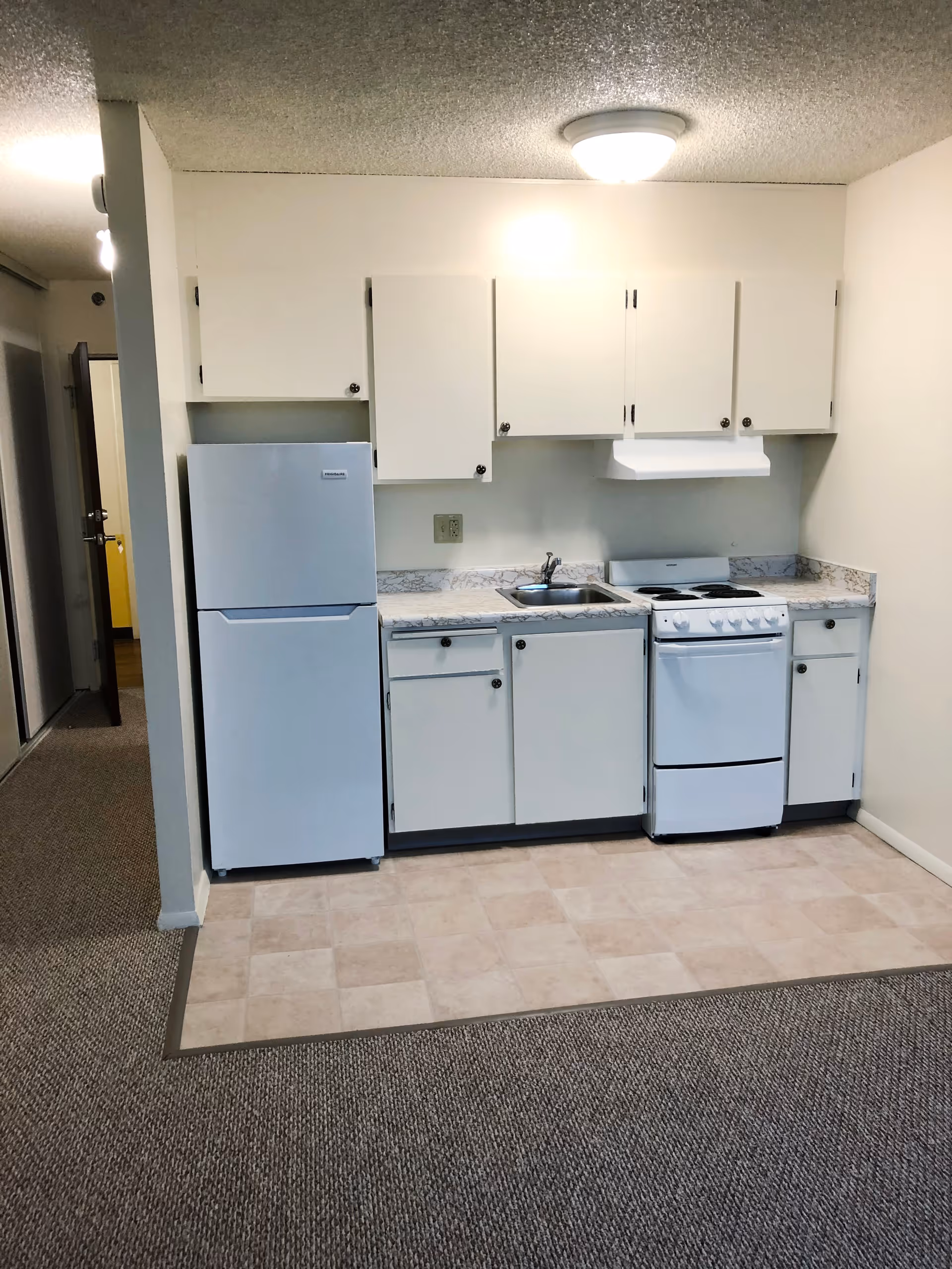 Small kitchen area with white cabinets, a white refrigerator, a white stove with oven, a stainless steel sink, and a marble-patterned countertop. The kitchen floor is tiled, and the surrounding area is carpeted. There is a ceiling light fixture above the kitchen.