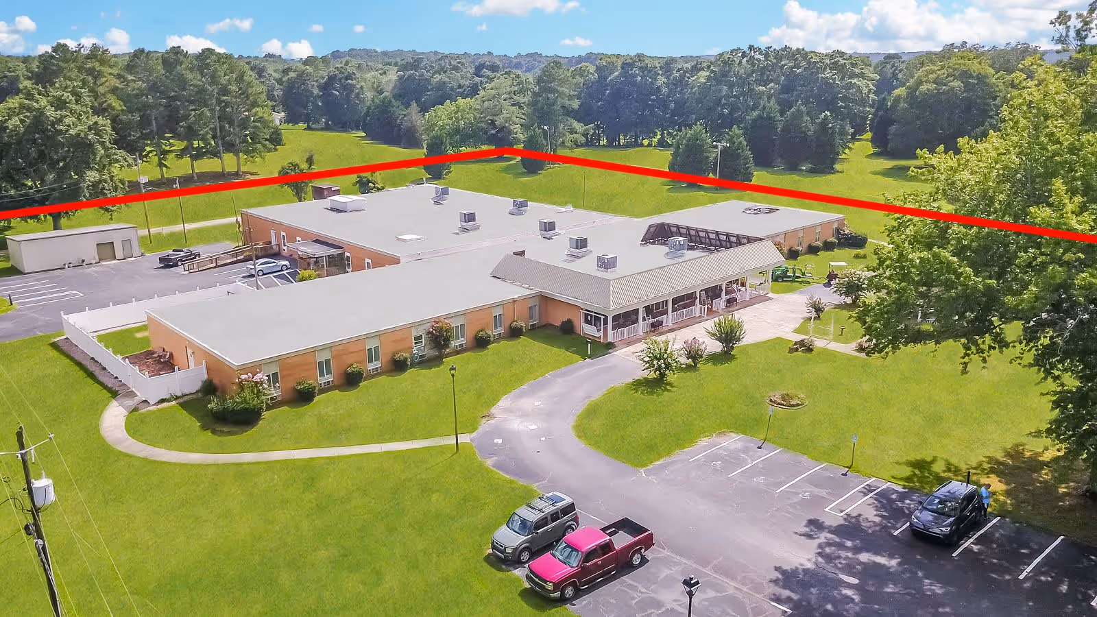 Aerial view of Front Porch of Bowdon Senior Living facility showing a single-story building with a flat roof surrounded by green lawns and trees. There is a parking lot with a few vehicles parked, and a paved driveway leading to the entrance. The background features a wooded area under a partly cloudy sky.