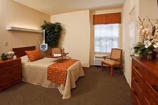 A furnished senior living bedroom with a bed, dresser, chair, window, and plants.
