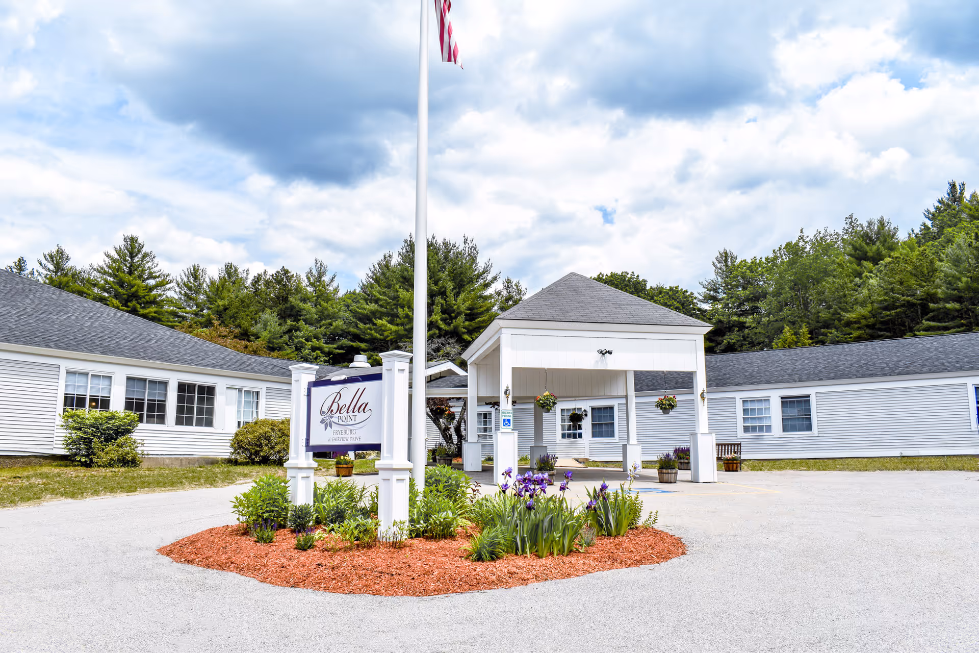 Front entrance of Bella Point Fryeburg with a sign, flagpole, covered porte-cochere and a landscaped flower bed with purple flowers.