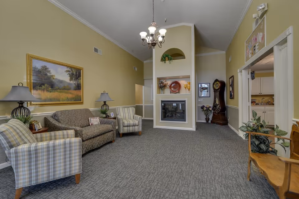 A cozy senior living facility common area with beige walls and gray carpet. The room features a patterned sofa and two striped armchairs arranged around a small wooden side table with lamps. A large framed landscape painting hangs on the wall. In the center, there is a built-in fireplace with decorative plates and vases on the mantel. A grandfather clock and a floral arrangement are visible in the background near an open doorway leading to a kitchen area. A chandelier hangs from the ceiling, and a wooden bench with a potted plant is on the right side.