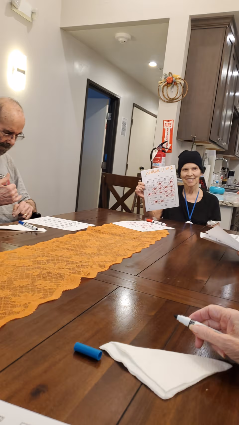 A group of seniors sitting around a wooden table playing bingo. One woman is smiling and holding up a bingo card marked with red Xs and the word 'WINNER!!' written at the top. The table has an orange lace runner, bingo cards, and markers. The background shows a kitchen area with cabinets and a fire extinguisher on the wall.