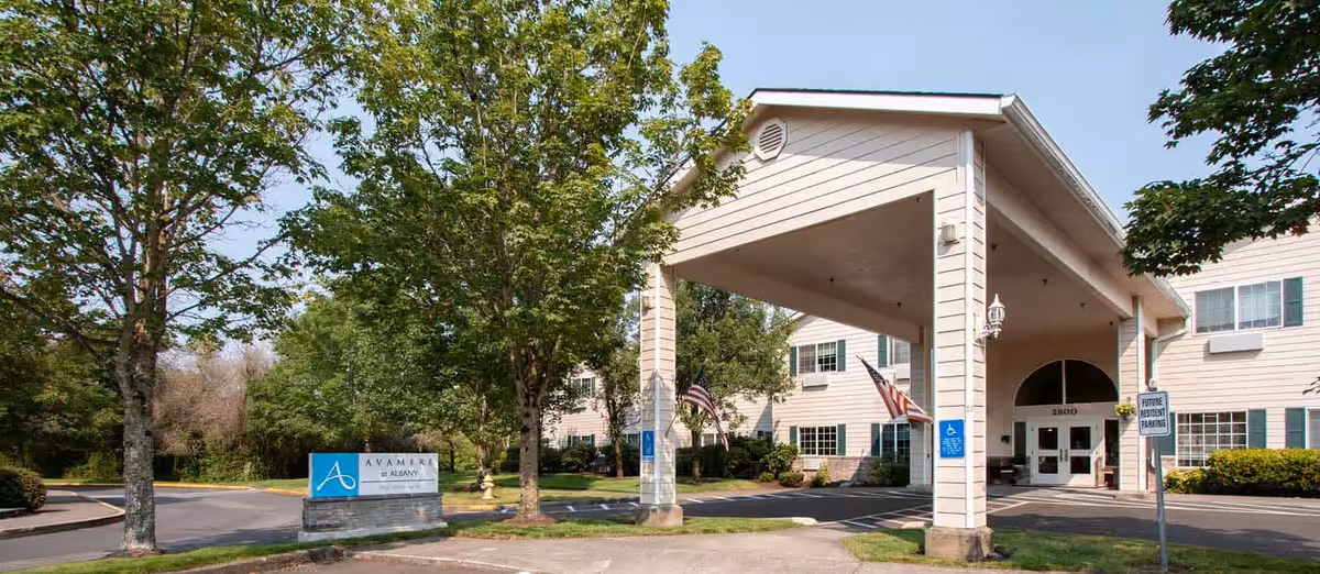 Exterior view of a senior living facility with a covered entrance, American flags, trees, and a sign that reads Avamere at Albany.