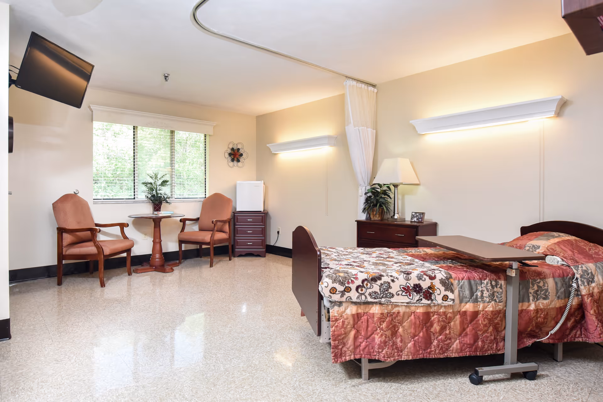 A well-lit senior living bedroom with a single bed covered in a floral patterned quilt. There is an overbed table on wheels positioned over the bed. Two upholstered chairs and a small round table are placed near a window with blinds. A flat-screen TV is mounted on the wall, and there are two dressers with a lamp, a plant, and a small photo frame. The room has light-colored walls and a tiled floor.