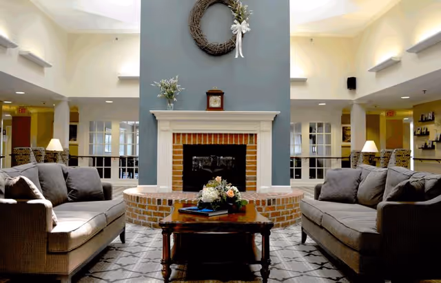 A cozy living room area with two gray sofas facing each other across a wooden coffee table with a floral centerpiece. Behind the table is a brick fireplace with a white mantel, decorated with a clock and a wreath above it. The room is well-lit with lamps and natural light from windows.