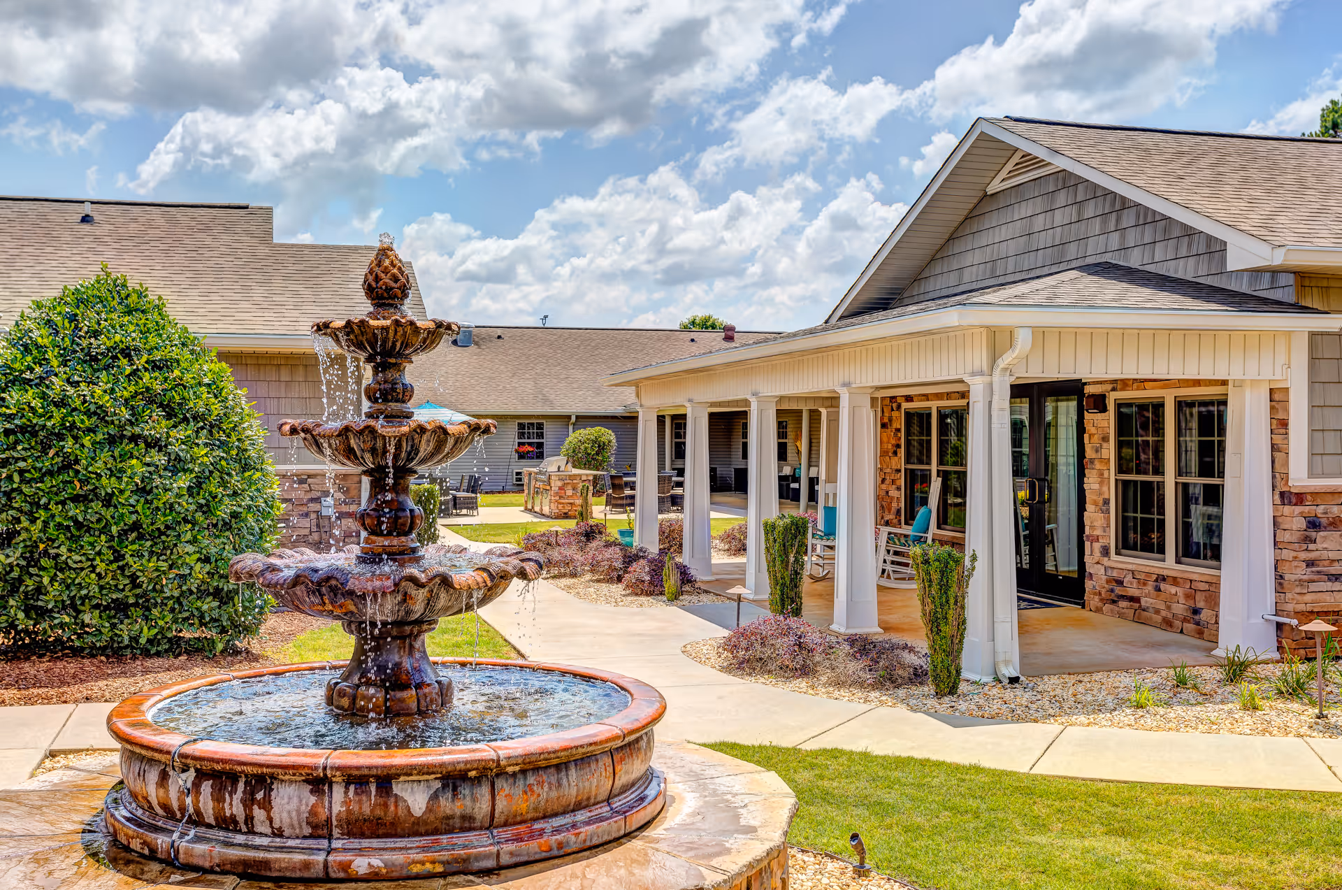 Outdoor view of a senior living facility courtyard with a multi-tiered water fountain in the foreground, surrounded by manicured landscaping and a paved walkway. The building features a covered porch with white columns, large windows, and a mix of brick and siding exterior under a partly cloudy sky.