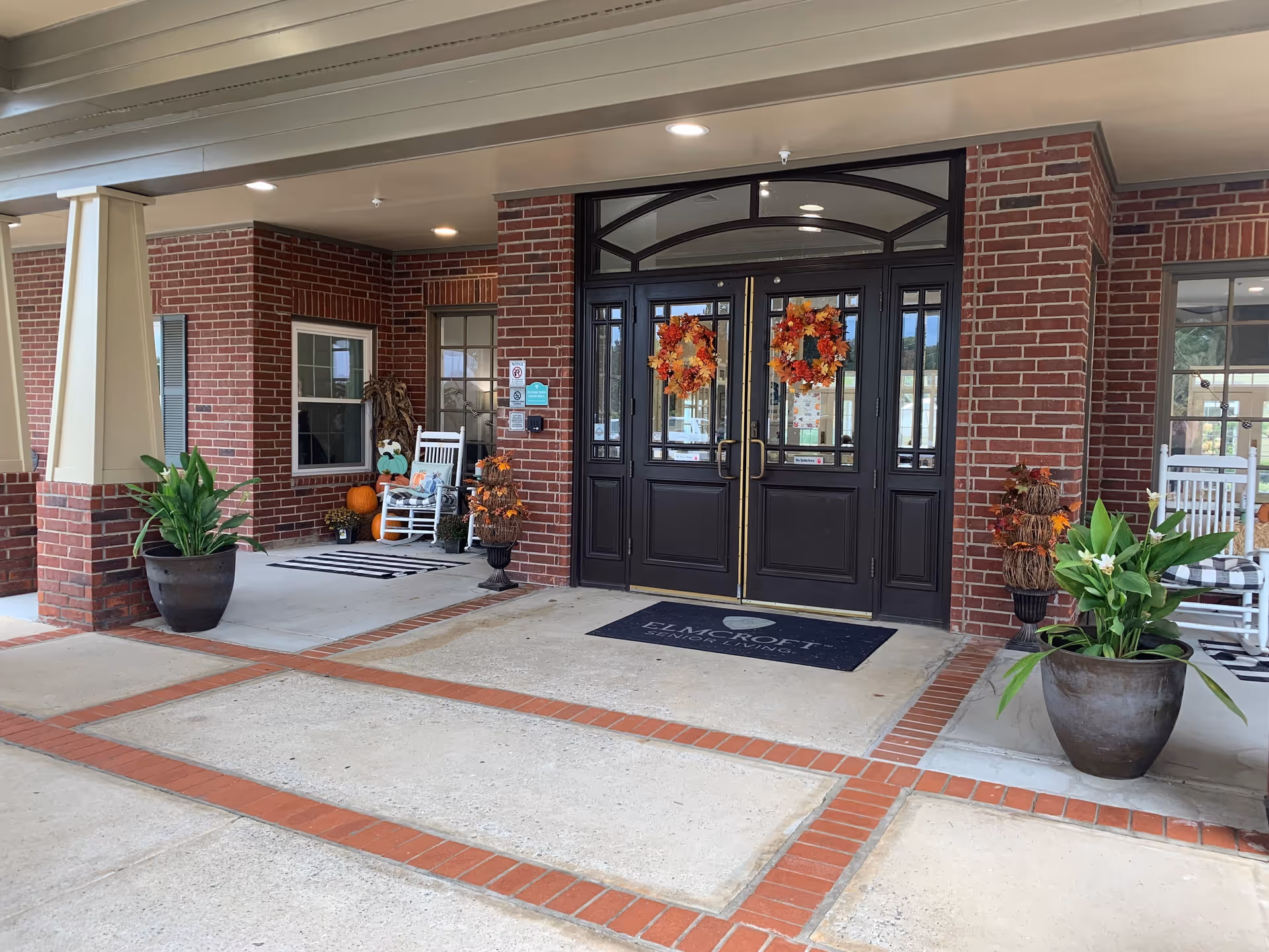 Entrance to a senior living facility with double black doors decorated with autumn wreaths. The building has red brick walls and a covered porch area with potted plants and white rocking chairs. Seasonal decorations including pumpkins and fall foliage are placed near the entrance.