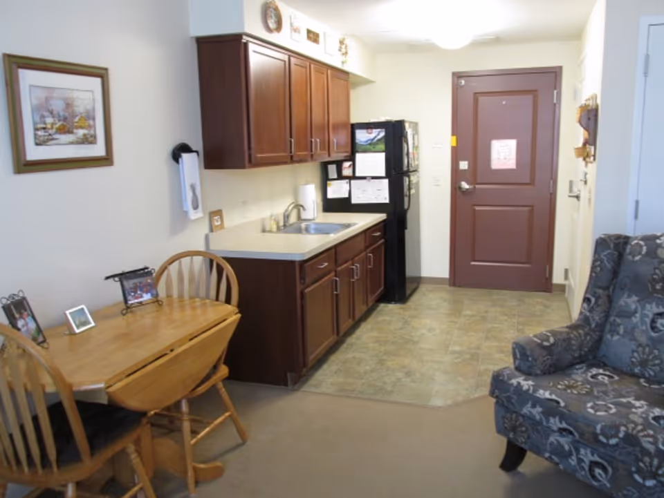 Small assisted living studio interior showing a kitchenette with dark wood cabinets, a dining table, upholstered armchair, and an entry door.