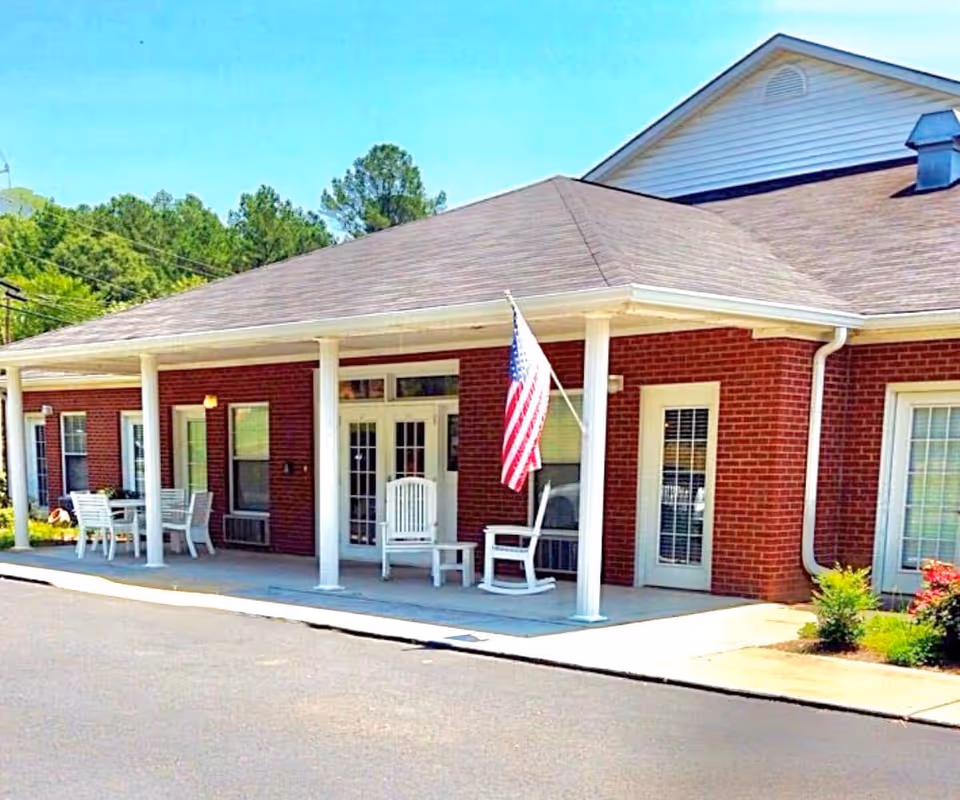 Exterior view of a single-story brick building with a covered porch featuring white columns. On the porch, there are white rocking chairs and a table with chairs. An American flag is mounted on one of the columns. The building has multiple windows and doors with white frames, and there is greenery around the building.