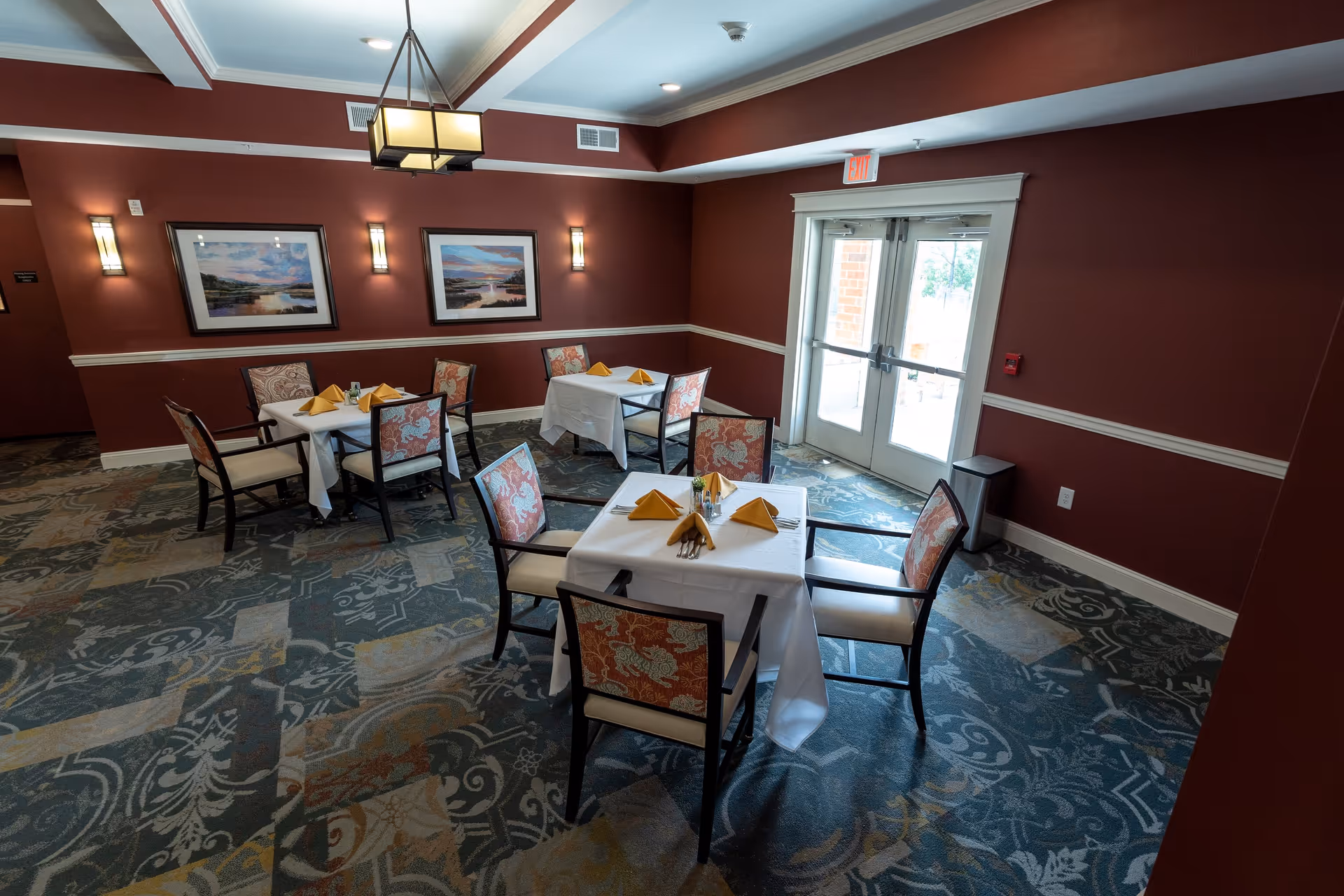 A dining room with three tables covered in white tablecloths, each set with yellow folded napkins and chairs with floral upholstery. The room has maroon walls with white trim, two framed landscape paintings, wall sconces, a patterned carpet, and a double glass door letting in natural light.