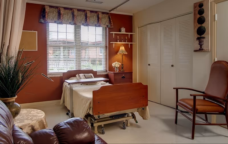 A cozy private bedroom with an adjustable medical bed, nightstand and lamp, seating, and a window with blinds and valance.