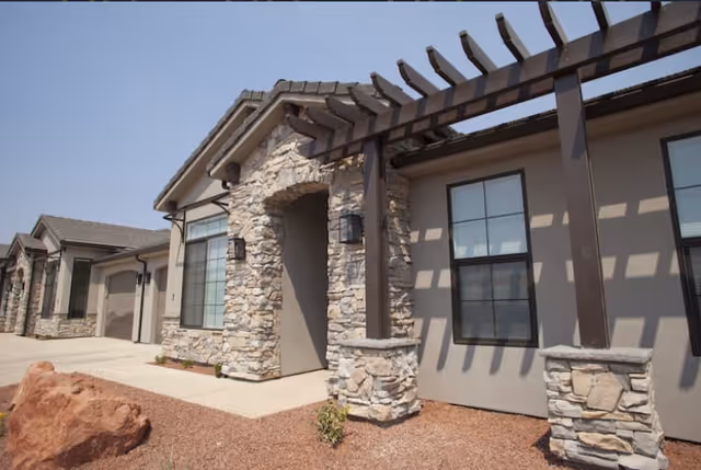 Exterior view of a senior living facility building with stone and stucco facade, large windows, and a pergola structure casting shadows on the wall. The ground is covered with reddish gravel and a large rock is visible in the foreground.
