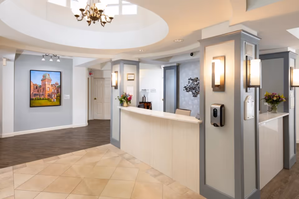Reception area of a senior living facility with a curved white reception desk, two vases with flowers, wall-mounted lights, a hand sanitizer dispenser, and a painting of a building on the wall. The floor is tiled near the desk and wood-floored further back, with a chandelier hanging from a circular ceiling feature.