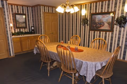 Dining room with a table covered by a tablecloth surrounded by six wooden chairs, a chandelier overhead, and striped wallpaper with framed art.