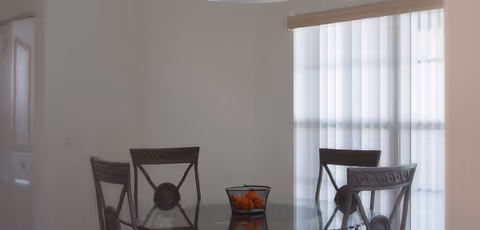 Glass dining table with four chairs and a bowl of oranges in a bright dining area.