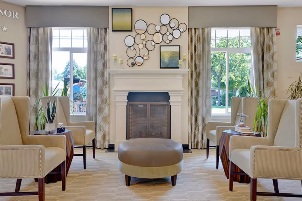 Bright lounge area with a central ottoman, upholstered armchairs facing a fireplace, and large windows with curtains.