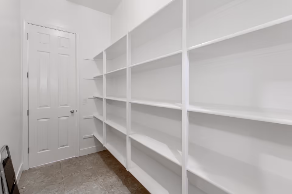 A narrow room with white built-in shelves lining the right wall and a closed white door at the end. The floor is covered with gray tiles, and the walls are painted white.