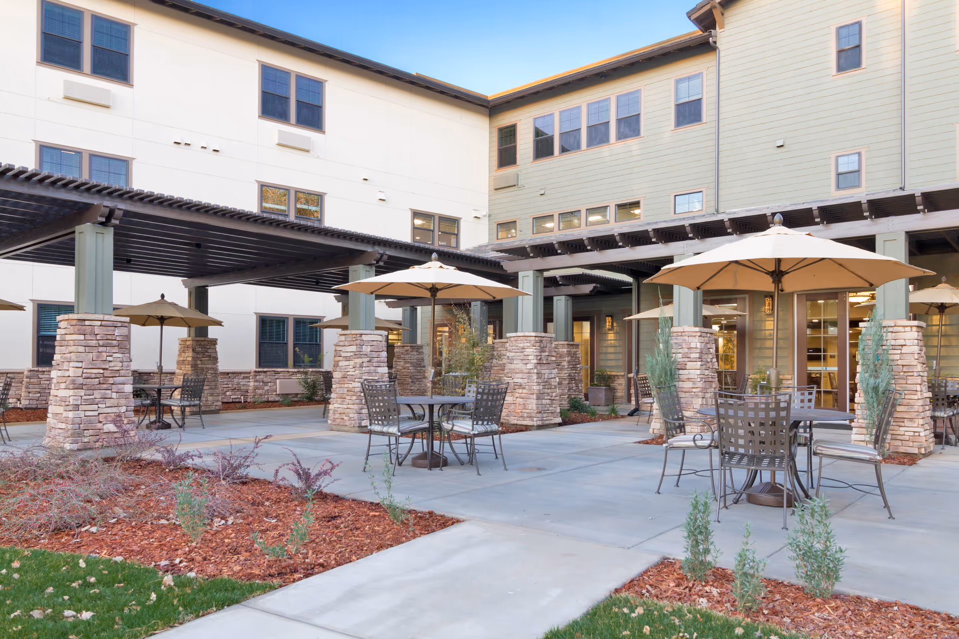 Outdoor patio area at Westmont of Chico with several round metal tables and chairs, each shaded by large beige umbrellas. The patio is surrounded by a multi-story building with light-colored walls and multiple windows. Stone pillars support pergolas over parts of the patio, and there are landscaped areas with mulch and small plants.