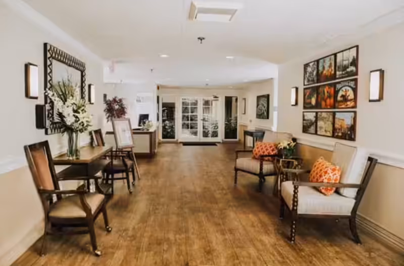 A well-lit hallway in an assisted living facility with wooden flooring, two cushioned armchairs with orange patterned pillows on the right, a small table with a flower vase and chairs on the left, framed artwork on the walls, and glass double doors at the far end.