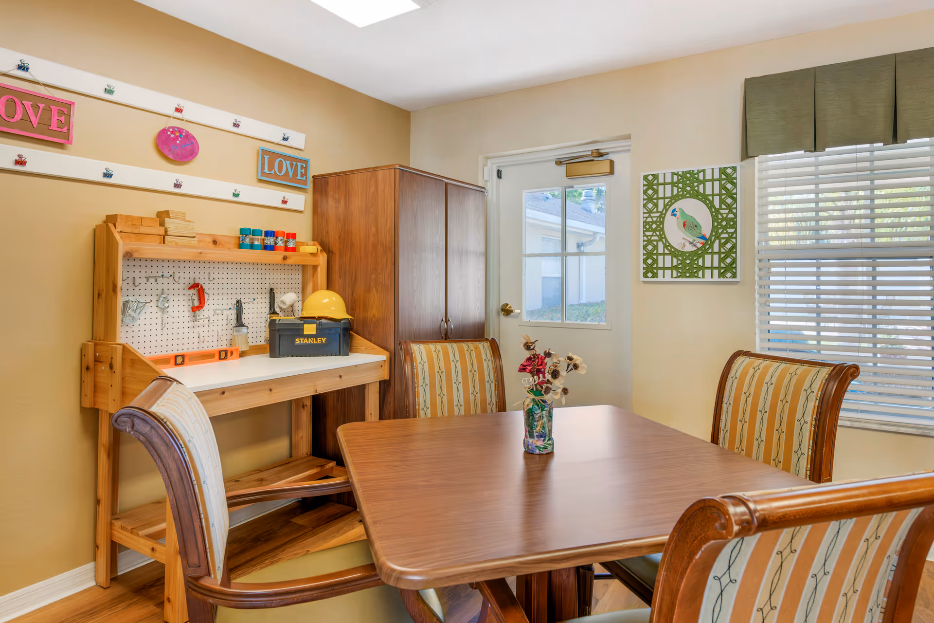 A cozy room with a wooden table and four upholstered chairs arranged around it. On the table is a small vase with decorative flowers. Against the wall is a wooden workbench with various tools, a yellow hard hat, and a Stanley toolbox. Above the workbench are two signs with the word 'LOVE' in pink and blue. A window with blinds and a door with a window are visible, along with a framed picture of a bird on the wall.