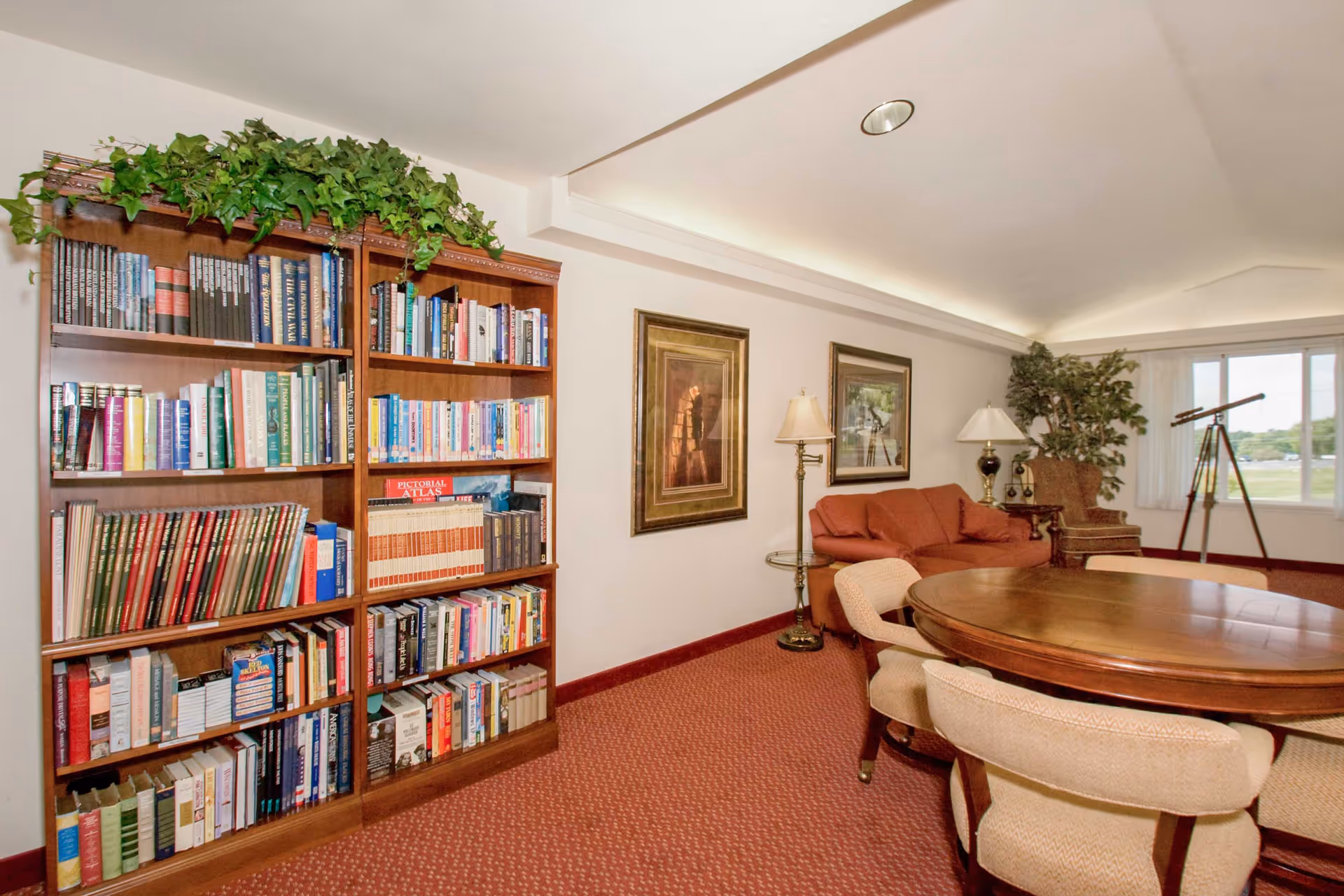A cozy senior living room with a wooden bookshelf filled with books and a green plant on top, a round wooden table surrounded by beige upholstered chairs, a red sofa, two framed paintings on the wall, two floor lamps, a large window with white curtains, a telescope, and a potted plant in the corner.