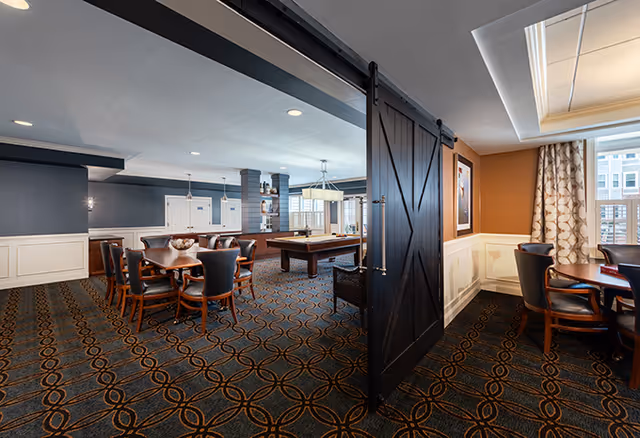 Interior view of a senior living facility's common area featuring a large room with a patterned carpet, multiple wooden tables and chairs, a pool table, and a sliding barn door separating two sections of the room. The walls are painted in dark blue and orange tones with white wainscoting, and large windows with curtains allow natural light to enter.