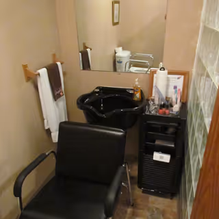 Small hair washing station with a black salon chair in front of a black sink. A large mirror is mounted on the wall behind the sink. To the right of the sink is a small black storage cart holding various hair care items and paper towels. A towel rack with white and brown towels is mounted on the left wall.