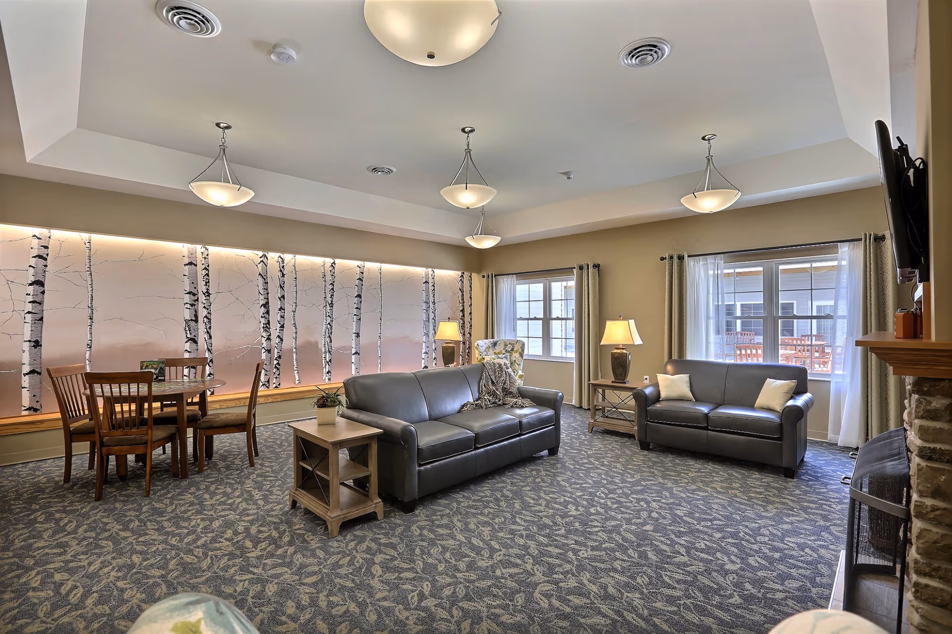 A bright communal living room with leather sofas, side tables, a dining set, a birch-tree wall mural, and multiple pendant lights.