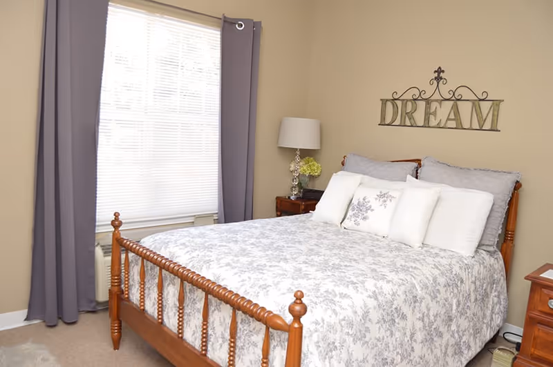 Sunlit bedroom with a wooden bed dressed in a floral bedspread and pillows, nightstands with a lamp and a window with gray curtains, and a 'DREAM' wall sign.